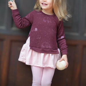 May include: A burgundy long-sleeved top with a light pink skirt and leggings. The top features a repeating elephant pattern and small white dots. The skirt has a ruffled hem. The child is holding a wooden ball.