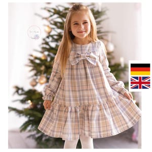Puede incluir: Un vestido a cuadros de color claro con mangas largas y un gran lazo en el escote. El vestido tiene un dobladillo con volantes y lo lleva una niña. Un árbol de Navidad es visible en el fondo.