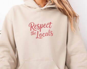 May include: A beige hooded sweatshirt with a front pocket and hood. The text "Respect the Locals" is printed in pink cursive. The person is wearing light blue jeans. The image is framed in a pink border.