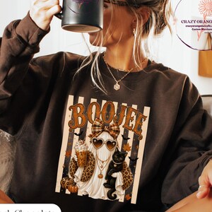 May include: A dark chocolate brown crewneck sweatshirt featuring a graphic print of a ghost wearing heart-shaped sunglasses, a plaid bow, and leopard print accents, holding a coffee cup and a black cat, with the text BOOJEE above the ghost.