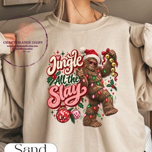 May include: A sand-colored crewneck sweatshirt featuring a graphic of a brown Bigfoot wearing a Santa hat and sunglasses, wrapped in colorful Christmas lights. The text reads Jingle All the Slay, accompanied by illustrations of holly, bells, and a disco ball ornament.