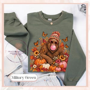 May include: A military green long-sleeve sweatshirt featuring a graphic of a brown sasquatch wearing a leopard print beanie and heart-shaped sunglasses while blowing a pink bubble. The design includes orange butterflies, sunflowers, and pumpkins.