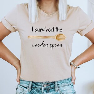 May include: A beige t-shirt with the text "I survived the wooden spoon" and a drawing of a wooden spoon.