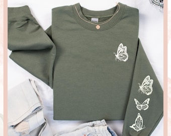 May include: An olive green sweatshirt with white butterfly designs on the chest and sleeve. The sweatshirt is paired with light-wash jeans. A gold necklace is visible at the neckline.