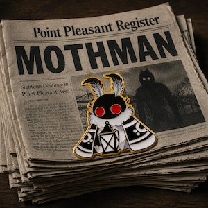 May include: A Mothman enamel pin with a gold outline, black body, red eyes, and white accents, on a stack of newspapers. The newspaper headline reads "MOTHMAN" and "Point Pleasant Register."