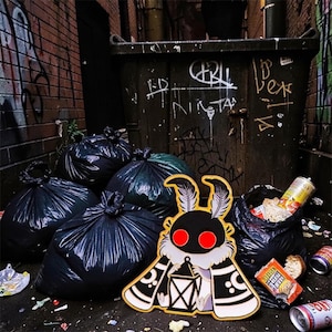 May include: A black and white moth pin with red eyes and a lantern design. The pin is set against a backdrop of black bin bags, a dumpster, and graffiti-covered brick walls, creating a unique contrast.
