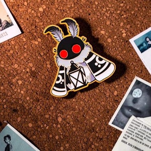 May include: A gold-coloured moth pin with a black body, red eyes, and white accents. The moth is holding a lantern and has feather-like antennae. The pin is on a brown surface with several small photographs scattered around it.