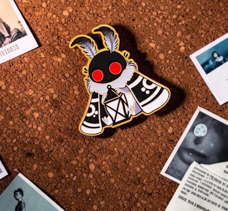 May include: A gold-coloured moth pin with a black body, red eyes, and white accents. The moth is holding a lantern and has feather-like antennae. The pin is on a brown surface with several small photographs scattered around it.