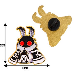 May include: A moth-shaped enamel pin, 3.2 cm high and 3.1 cm wide, featuring a black head, red eyes, and a white and black patterned cape. The pin's reverse is gold with a black pin clasp.