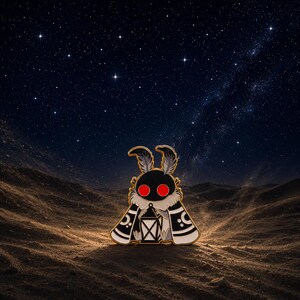 May include: A moth-shaped enamel pin with a black body, red eyes, and a lantern. The pin features gold edging and white details on its wings. The background shows a starry night sky over a desert landscape.