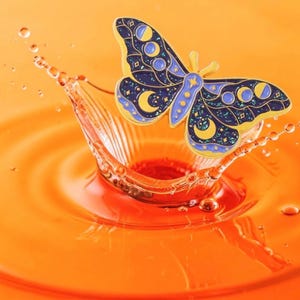 May include: A decorative butterfly pin with a celestial design. The pin features a navy blue body with yellow and blue accents, including crescent moons and stars. The pin is set against an orange background with water droplets.