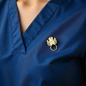 May include: A gold-colored angel pin with white and yellow enamel details and a red gem. The pin is attached to a dark blue garment.