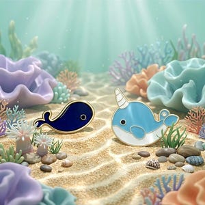 May include: Two whale-shaped enamel pins. One is a dark blue whale with a gold outline, and the other is a light blue narwhal with a white belly and a gold outline. The pins are set on a sandy ocean floor.