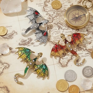 Pode incluir: Quatro broches de dragão em várias cores, incluindo verde, vermelho e prata, são exibidos em um mapa vintage. Cada broche apresenta detalhes intrincados e acentos brilhantes. Uma bússola e moedas também estão presentes.