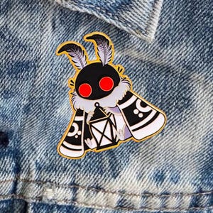 May include: A gold enamel pin depicting a stylized moth. The moth has a black head with red eyes, feathery antennae, and a white and black body holding a lantern. The pin is set against a denim background.
