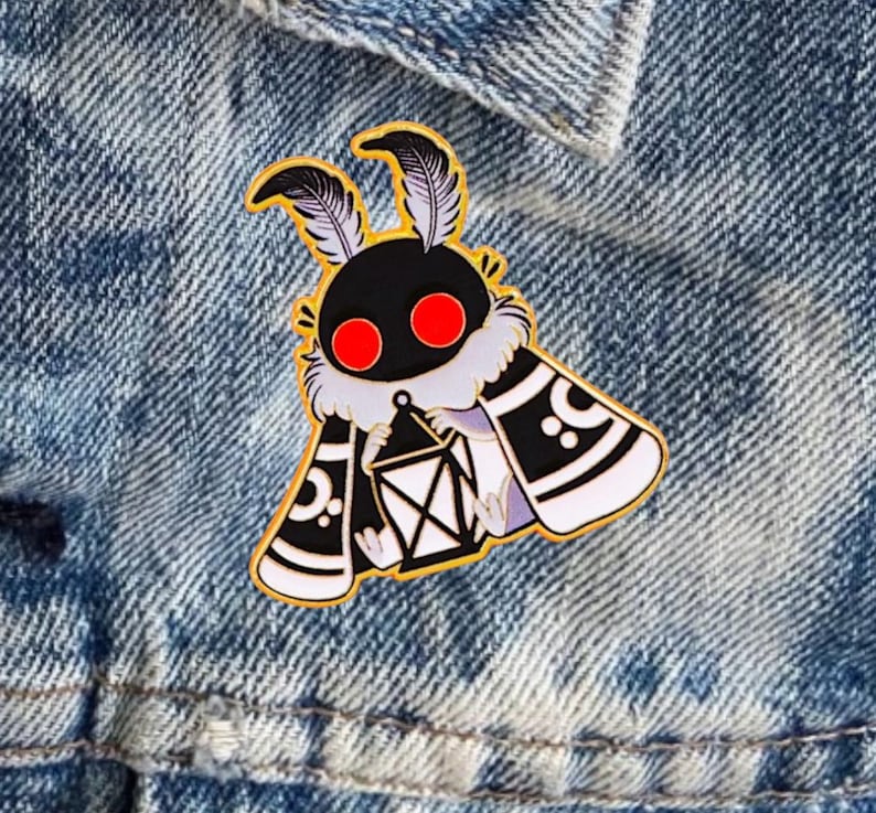May include: A gold enamel pin depicting a stylized moth. The moth has a black head with red eyes, feathery antennae, and a white and black body holding a lantern. The pin is set against a denim background.