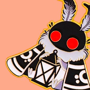 May include: A quirky enamel pin depicting a moth-like character with a black body, red eyes, and white details. The moth holds a lantern and has feathery antennae. The pin has a gold outline and is set against a peach background.
