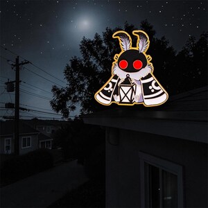 May include: A moth-shaped enamel pin with a black body, red eyes, and a lantern. The moth has white and black patterned wings, set against a night sky with stars and a full moon.