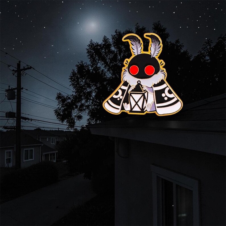 May include: A moth-shaped enamel pin with a black body, red eyes, and a lantern. The moth has white and black patterned wings, set against a night sky with stars and a full moon.
