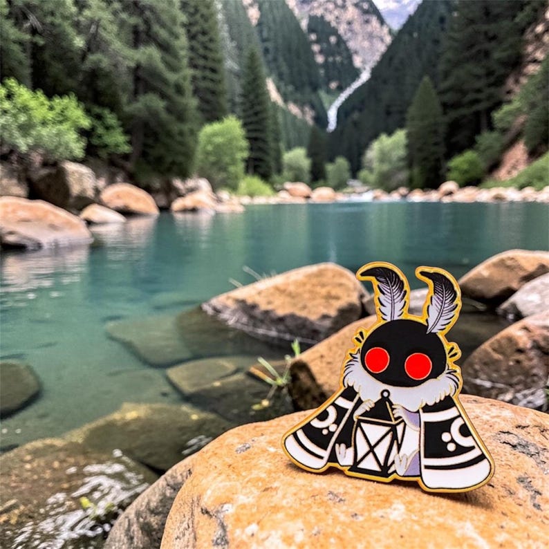 May include: A black and white moth enamel pin with a gold outline, red eyes, and a lantern design. The pin has a white fur-like collar. The pin is set against a scenic backdrop of a lake and mountains.