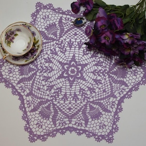 Puede incluir: Un encaje de ganchillo morado con un centro en forma de estrella y diseños florales intrincados. El encaje está colocado sobre una superficie blanca con una taza de té y un platillo y flores moradas.