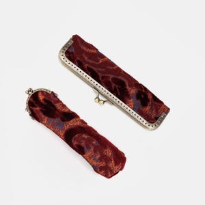 May include: Two elongated cases with a deep burgundy velvet texture and a floral pattern in orange and blue tones. Each case features a metallic frame with a kiss-lock clasp closure, finished in an antique brass tone with a perforated edge detail.