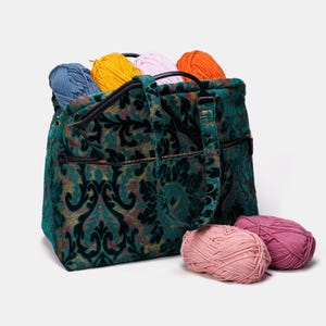 Puede incluir: Una bolsa de mano de terciopelo verde con un patrón de damasco, con una cremallera negra y una correa negra. La bolsa está llena de ovillos de lana de colores, incluyendo azul, naranja, amarillo y rosa.