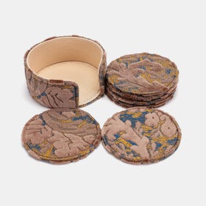 May include: A set of six round coasters with a matching holder. The coasters and holder are covered in a brown fabric with a floral pattern in blue and gold. The holder has a beige interior.