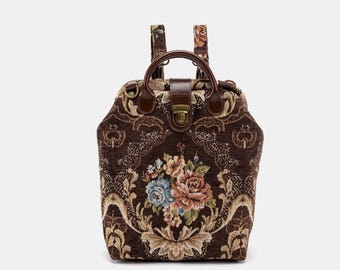 May include: A brown tapestry backpack with a floral design. The bag features a top handle, adjustable straps, and a clasp closure. The floral pattern includes roses and other flowers in shades of pink, blue, and beige. The bag is approximately 12 inches tall.