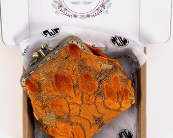 Puede incluir: Una pequeña cartera de monedas de terciopelo naranja con un patrón floral y un cierre de latón. La cartera está en una caja marrón con papel de seda blanco.