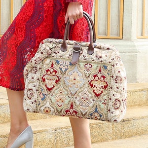 Puede incluir: Una gran bolsa de viaje de color crema con un asa de cuero marrón y un tejido estampado con intrincados motivos rojos, azules y dorados. La bolsa tiene un diseño tradicional y ornamentado.