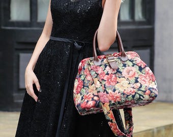 May include: A floral print handbag with brown leather handles and a matching shoulder strap. The bag features a colorful pattern of red, pink, and yellow flowers on a dark background. The bag is held by a person wearing a black dress.