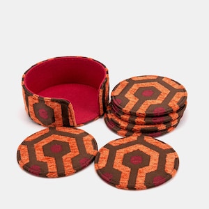 May include: A set of drink coasters with a matching holder. The coasters and holder feature a repeating geometric pattern in shades of orange, brown, and burgundy. The holder has a red interior.