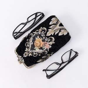 May include: A black eyeglass case with a floral design and two pairs of black-framed spectacles. The case has a clasp closure and a decorative floral pattern in gold, pink, and green.