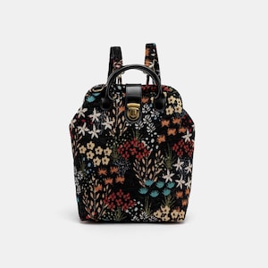 May include: A black backpack with a floral pattern. The backpack has a top flap with a gold buckle closure and adjustable straps.