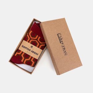 Puede incluir: Un par de calcetines de algodón burdeos con un diseño geométrico naranja, presentados en una caja de cartón marrón abierta. Los calcetines están etiquetados con "Cotton Socks" en una banda de papel marrón. La caja muestra el nombre de la marca "Carpetbag".