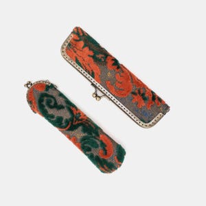 May include: Two elongated cases with a kiss-lock metal frame closure. The exterior features a textured fabric with a repeating pattern of orange and dark green floral motifs against a grey background. The metal hardware has a metallic finish.
