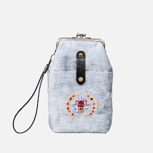 Op de afbeelding: Een kleine grijze fluwelen clutch met een zwarte band en een gouden sluiting. De tas heeft een geborduurd ontwerp van een rood en gouden dierengezicht met blauwe accenten.