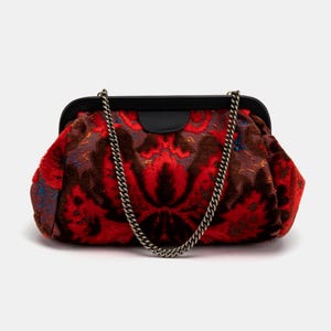 May include: A red and brown patterned clutch bag with a silver chain strap. The bag has a black frame and clasp. The pattern features a floral design. The clutch is a small, handheld accessory.