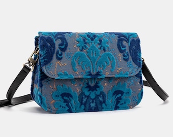 May include: A blue crossbody bag with a flap closure. The bag features a damask pattern in shades of blue and teal, with gold accents. It has a black adjustable strap and gold-tone hardware.
