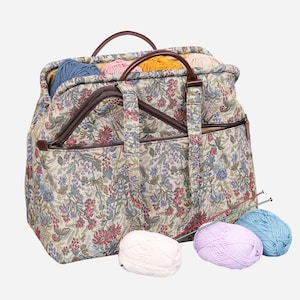 May include: A tapestry-style knitting bag with brown leather handles and multiple pockets. The bag is filled with balls of yarn in various colors, including pink, blue, and yellow. There are also knitting needles on the table next to the bag.
