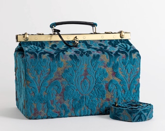 Könnte beinhalten: Eine Arzt-Tasche aus blaugrünem Samt mit goldenen Beschlägen und einem passenden Riemen. Die Tasche hat ein Damastmuster.