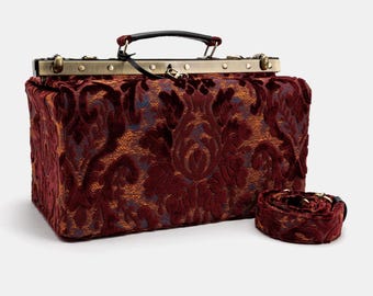 Könnte beinhalten: Eine bordeauxrote Samt-Arzttasche mit goldenem Metallrahmen und einem passenden Samtband. Die Tasche hat ein Blumenmuster.