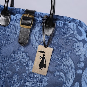 Peut inclure: Un sac cabas en velours bleu avec un motif floral. Le sac a une poignée en cuir noir et une boucle en cuir noir. Une étiquette brune avec une silhouette noire d'une femme tenant un parapluie est attachée au sac. L'étiquette indique "Mary Poppins".
