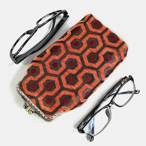 May include: A red and brown patterned glasses case with a metal clasp. The case is made of fabric and has a geometric design. Two pairs of black framed glasses are also pictured.