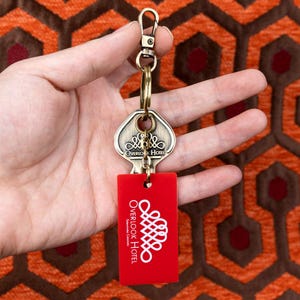 May include: A gold-coloured key chain with a key shaped like a hotel room key. The key has the words "Overlook Hotel" engraved on it. The keychain also has a red plastic tag with the words "Overlook Hotel" and "Snowden, Colorado" printed on it.