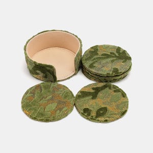 Puede incluir: Un juego de posavasos redondos con estampado verde y dorado con un soporte a juego. Los posavasos son de tela texturizada. El soporte es un recipiente redondo y abierto con interior beige.