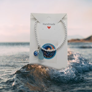 May include: A silver bracelet with a charm featuring a blue sea glass piece and a silver charm with a wave design. The bracelet is on a white card with the word "Handmade" and a red heart.