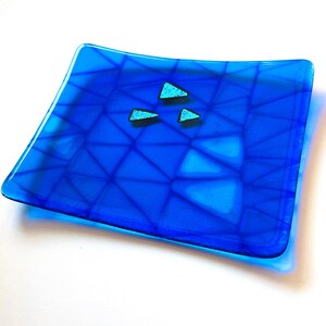 May include: A square, blue glass dish with a geometric pattern and three small, turquoise triangle accents. The dish has a glossy finish and is likely handmade.
