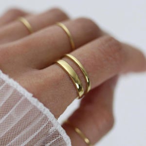 Könnte beinhalten: Nahaufnahme mehrerer goldener Ringe an einem Finger. Die Ringe sind in verschiedenen Stilen gehalten, darunter dünne Bänder und breitere, flache Bänder. Die Ringe sind glänzend und reflektieren das Licht, wodurch ein warmer, goldener Schein entsteht. Der Hintergrund ist unscharf, wodurch der Schmuck betont wird.